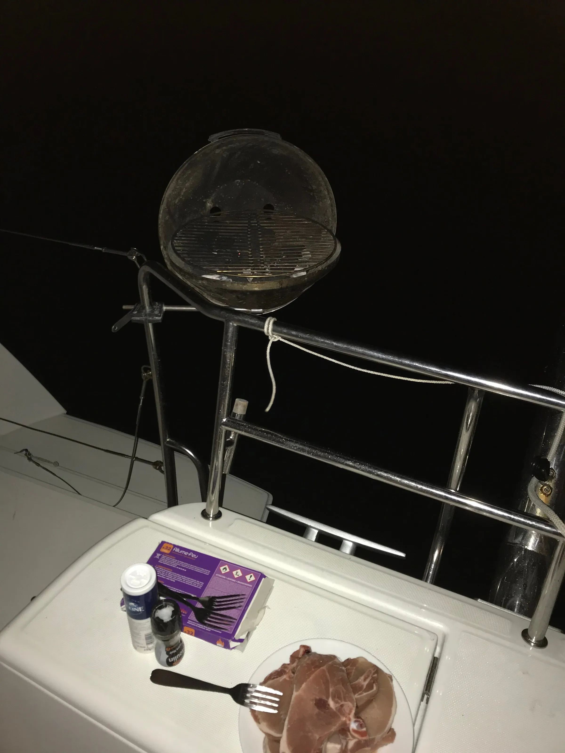 A plate of raw meat beside a small barbecue fixed to the catamaran's aft rail at night in the anchorage.