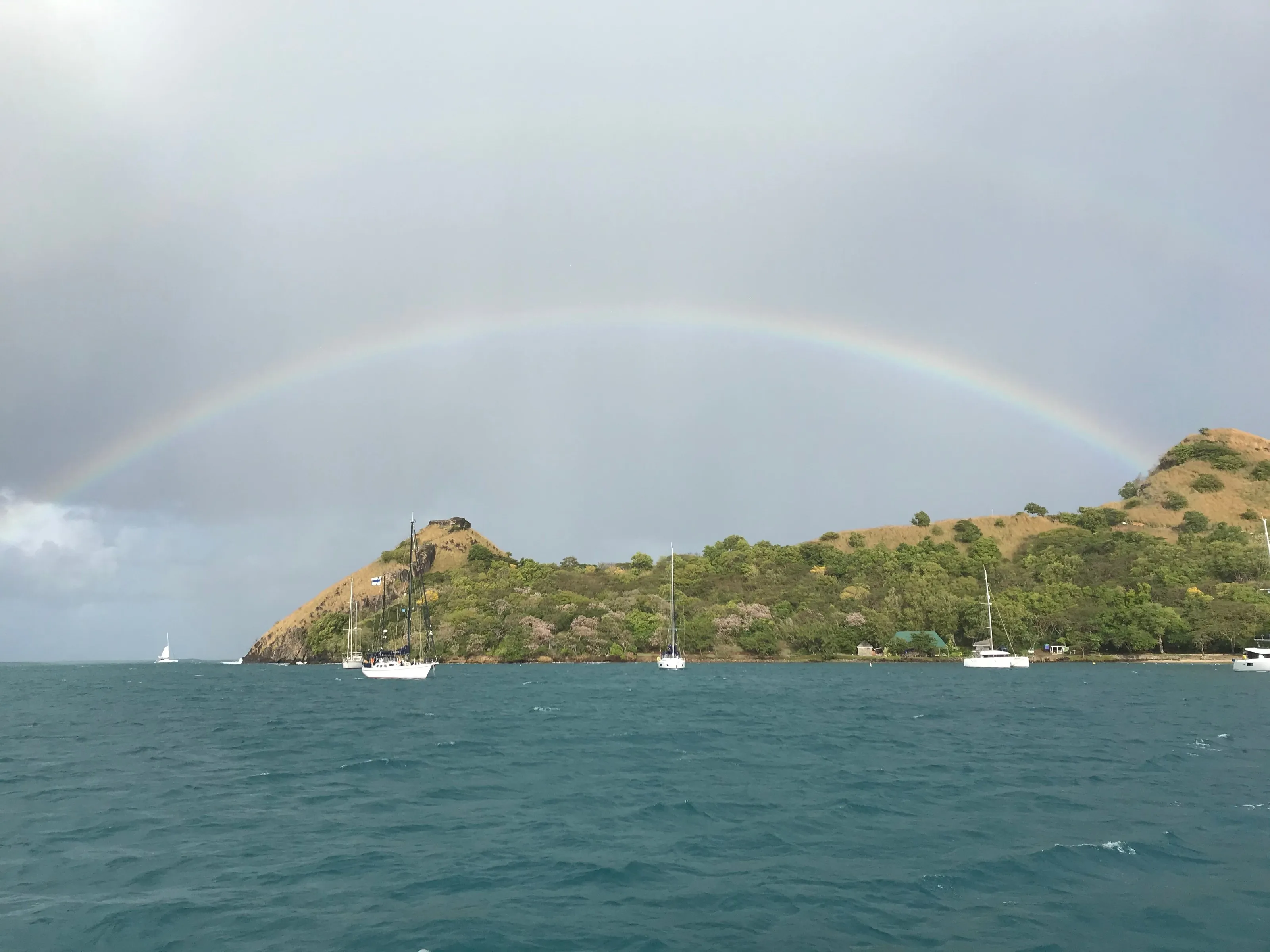 A rainbow above a green hill and yachts at anchor in Rodney Bay, Saint Lucia, beneath a rainy sky.