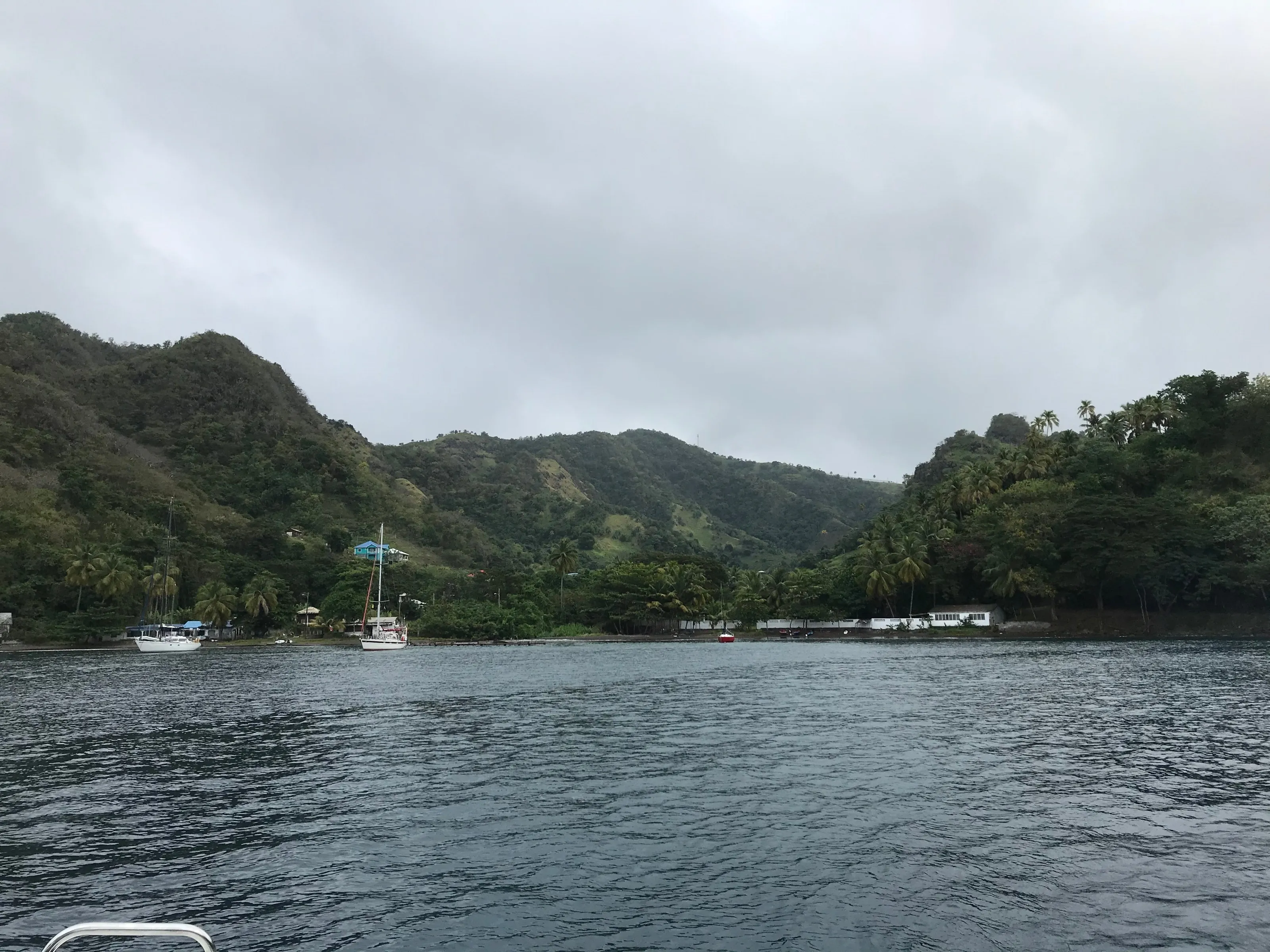 Wallilabou Bay on Saint Vincent, enclosed by green hills, with two yachts at anchor beneath a grey sky.