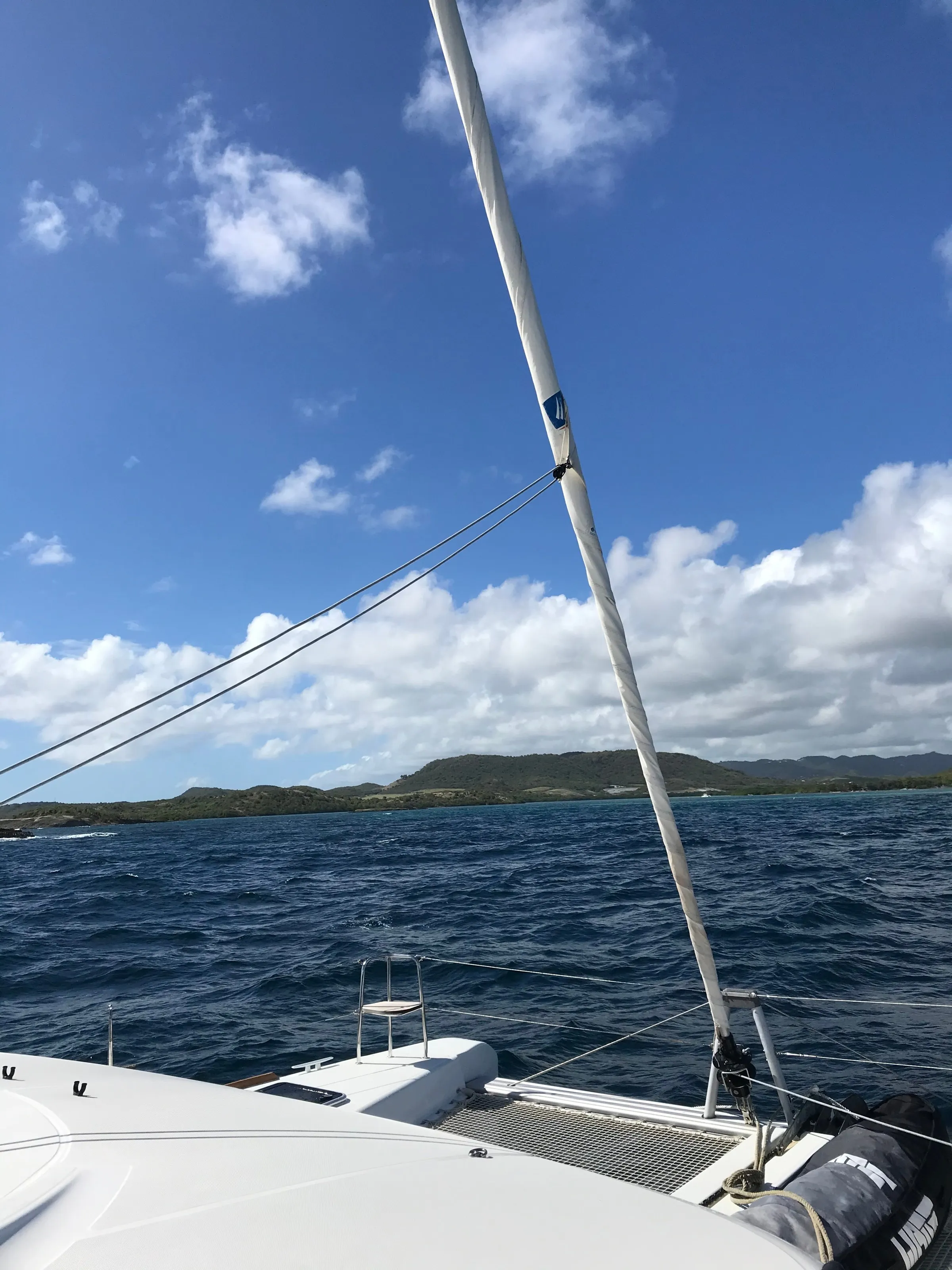 The catamaran's foredeck and furled sail facing a green coast beneath a blue sky crossed by clouds.