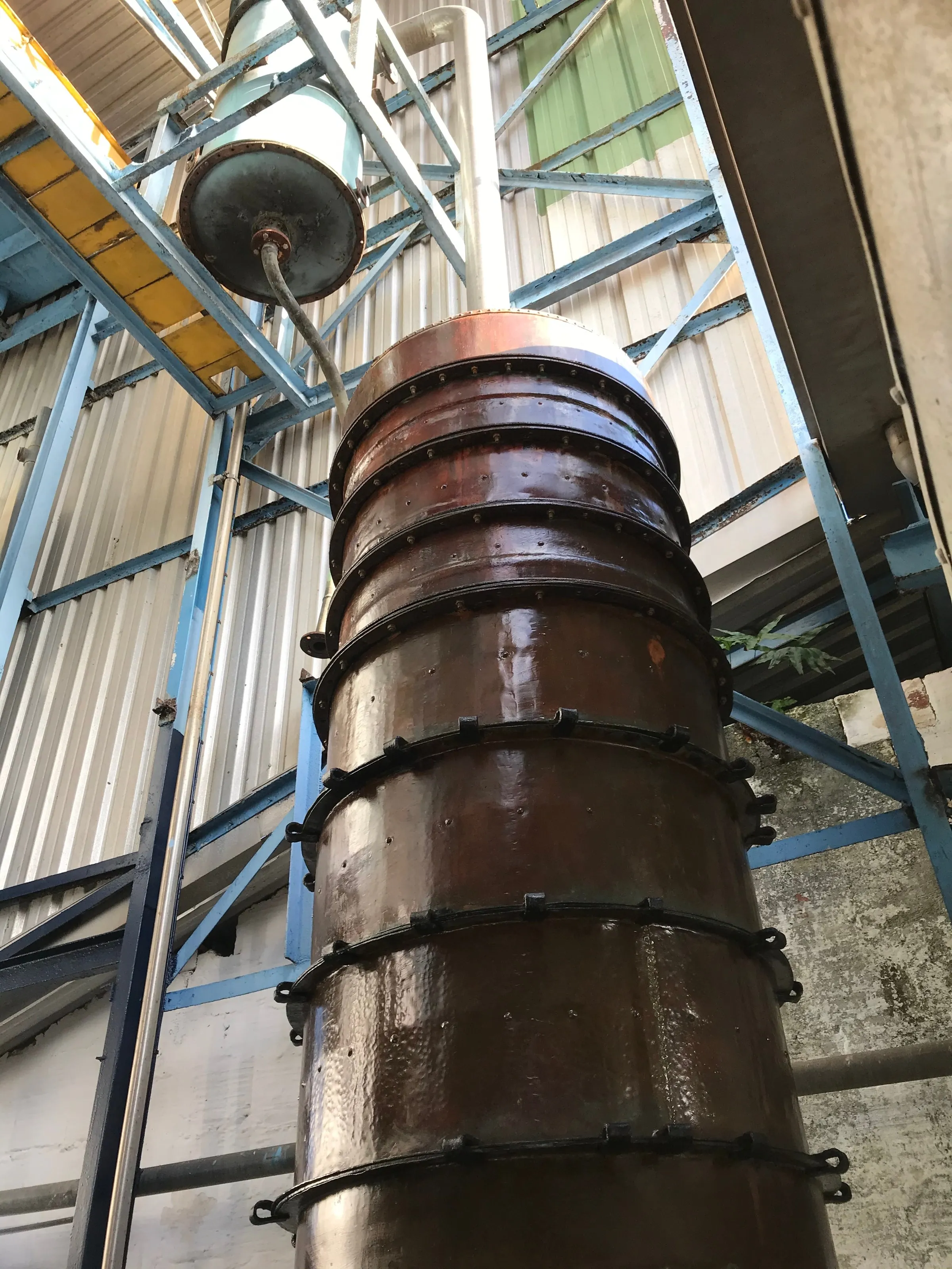 A copper distillation column seen from below beneath the metal roof structure of a distillery in Sainte-Luce, Martinique.