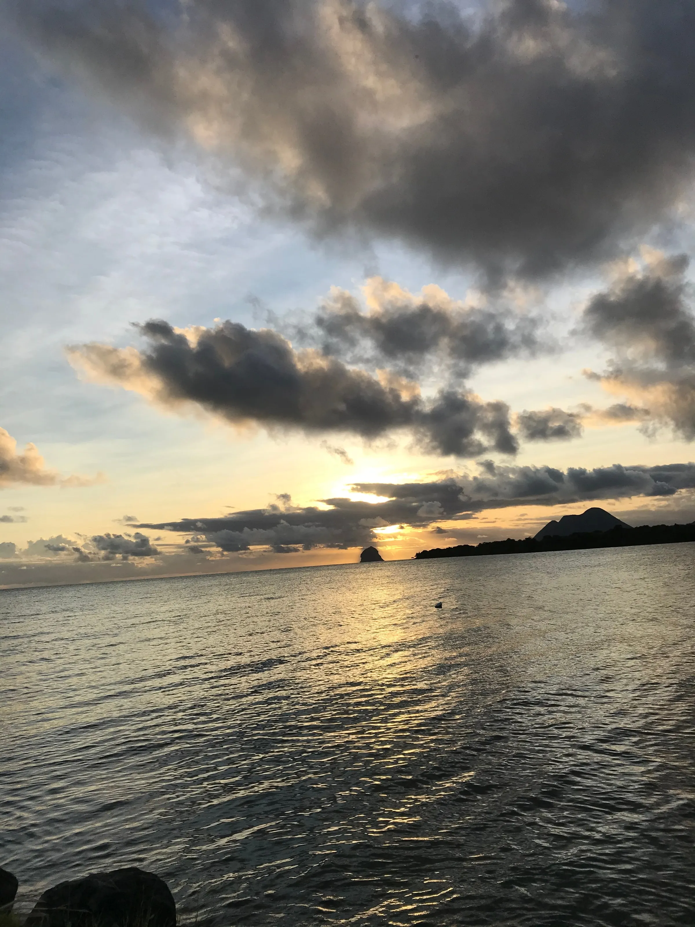 The sun setting between clouds behind an isolated rock, seen from the Sainte-Luce coast above a calm sea.