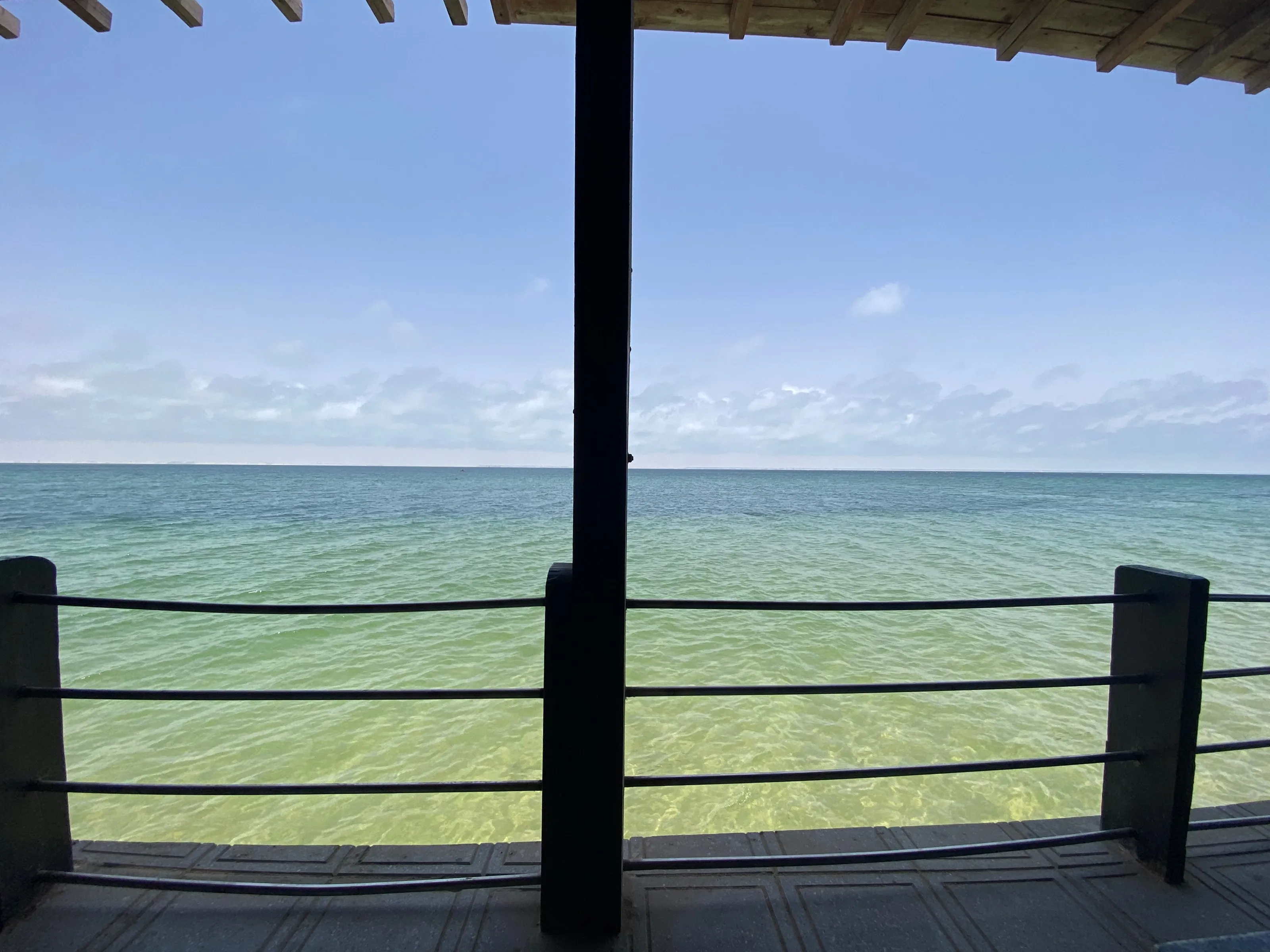The clear green water of Dakhla lagoon seen between the posts of a covered wooden terrace and beyond a black-cable railing.