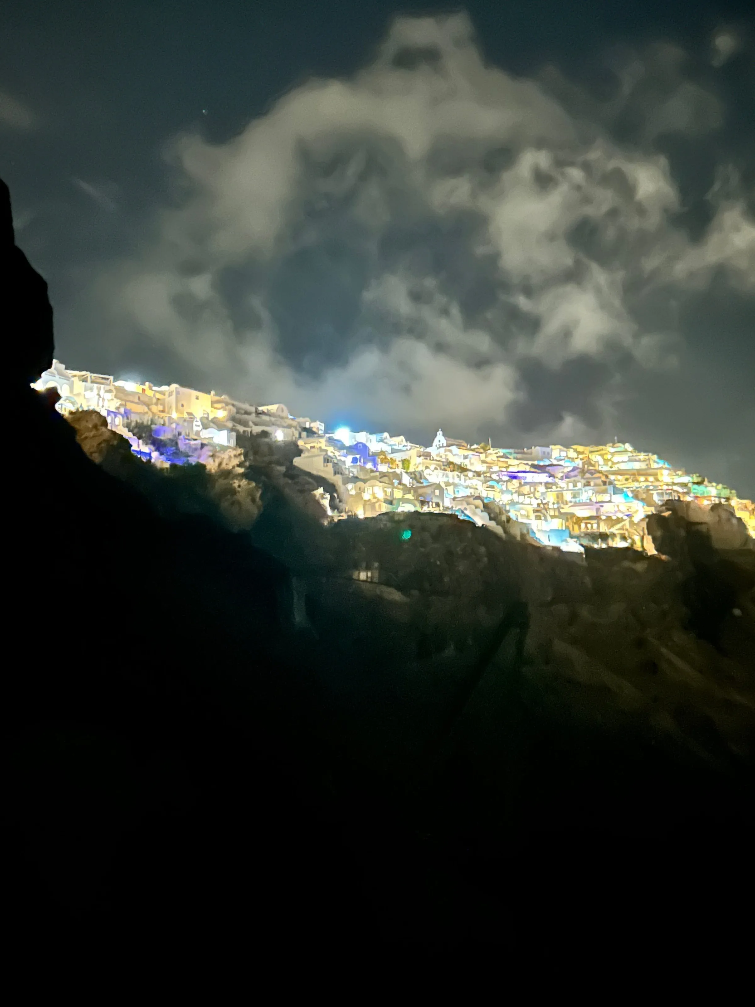 Oia, a white village on Santorini in the Cyclades, illuminated on top of a dark cliff beneath a cloudy night sky.