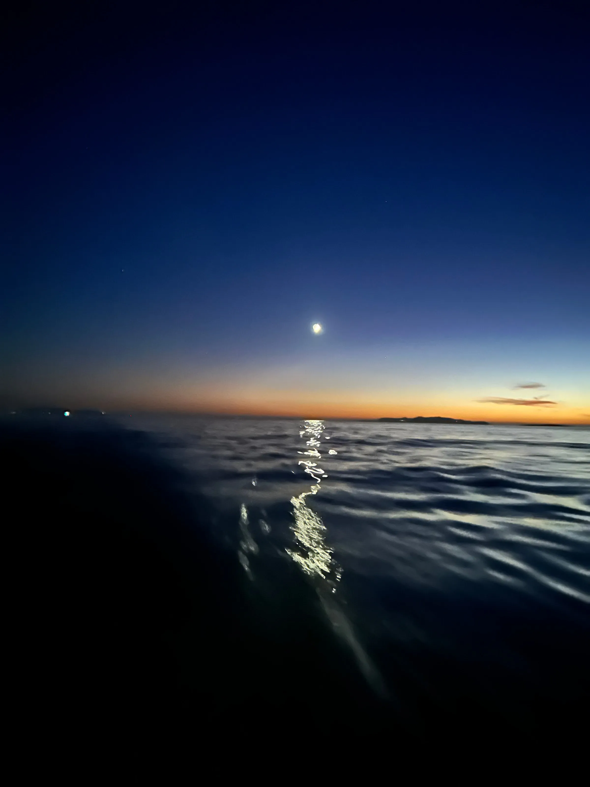 Moonlight reflected on rippled water beneath an orange and deep-blue dusk sky in Foinikas Bay, Syros, in the Cyclades.