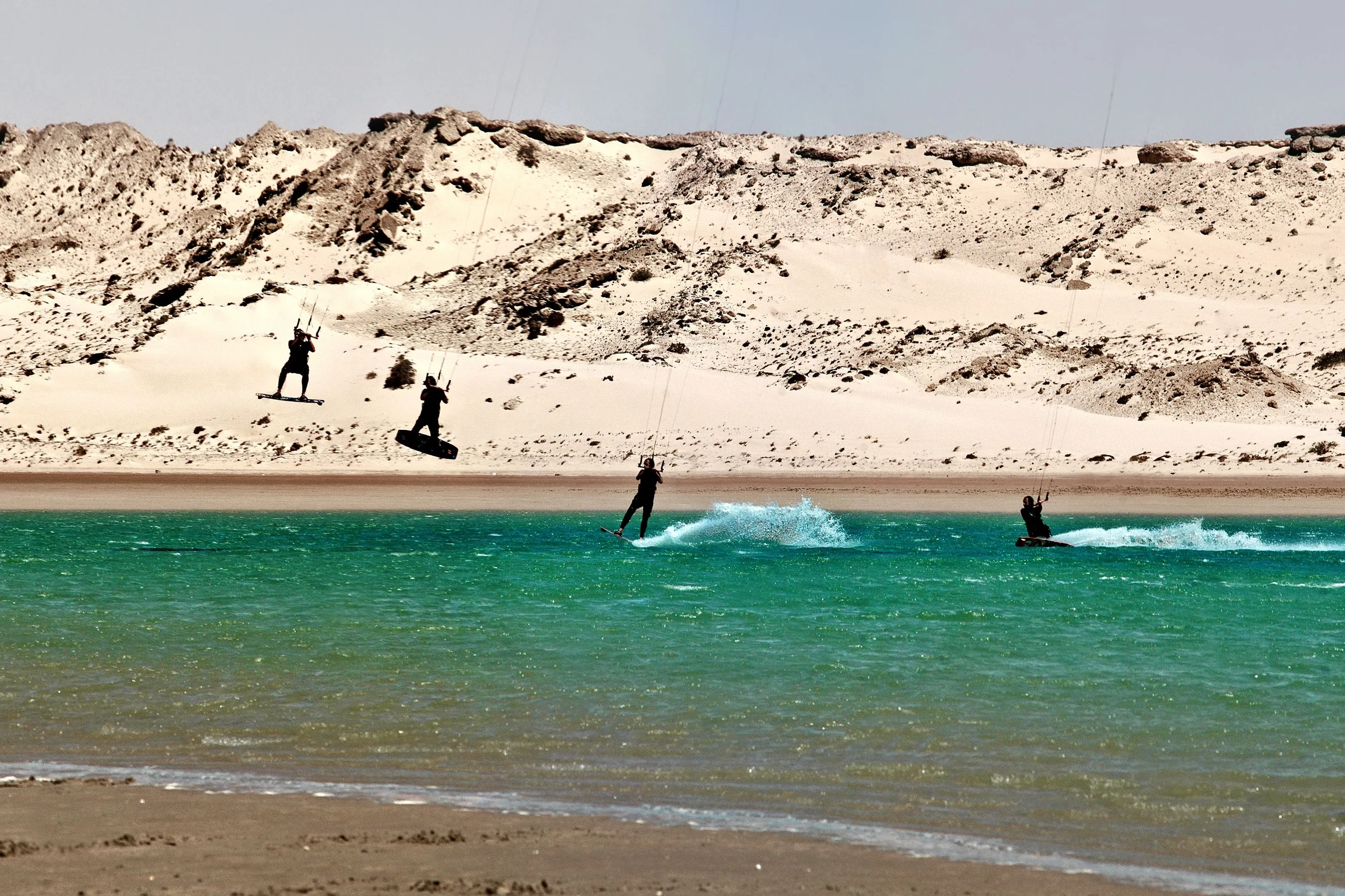 Four silhouetted kitesurfers, two of them airborne, on the green water of Dakhla lagoon in front of a pale dune.